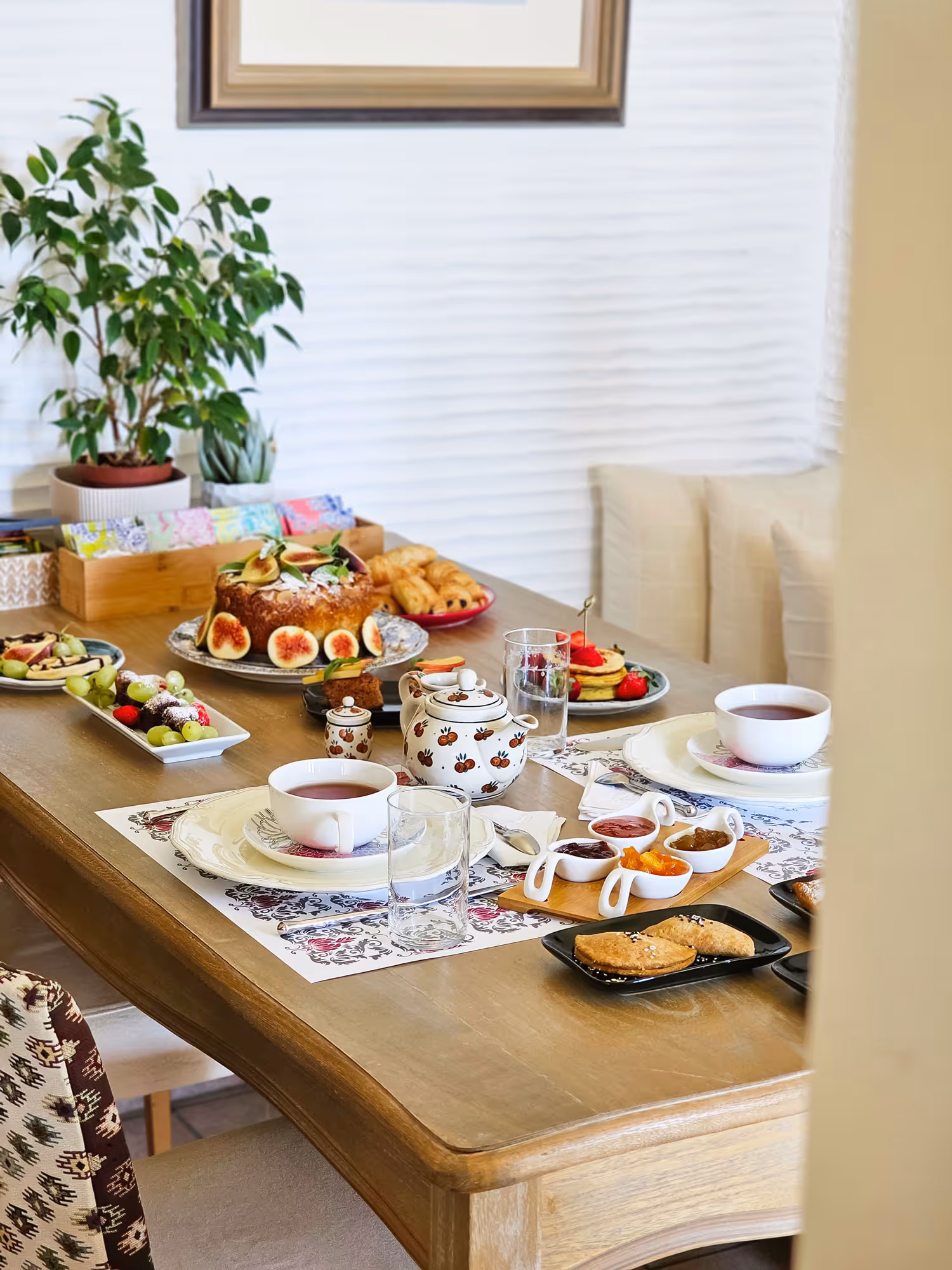 Table set for tea with cups, teapot, pastries, fruit, jams, and cake in a cozy dining room.
