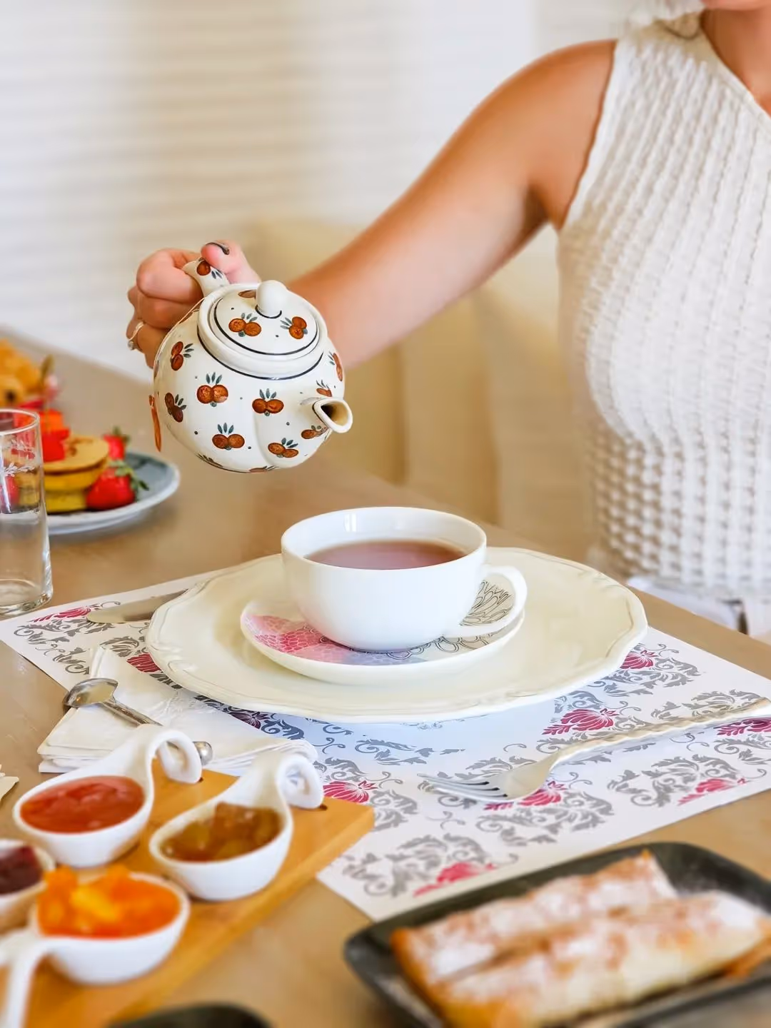 Person pouring tea from floral teapot into white cup on plate with breakfast condiments nearby.