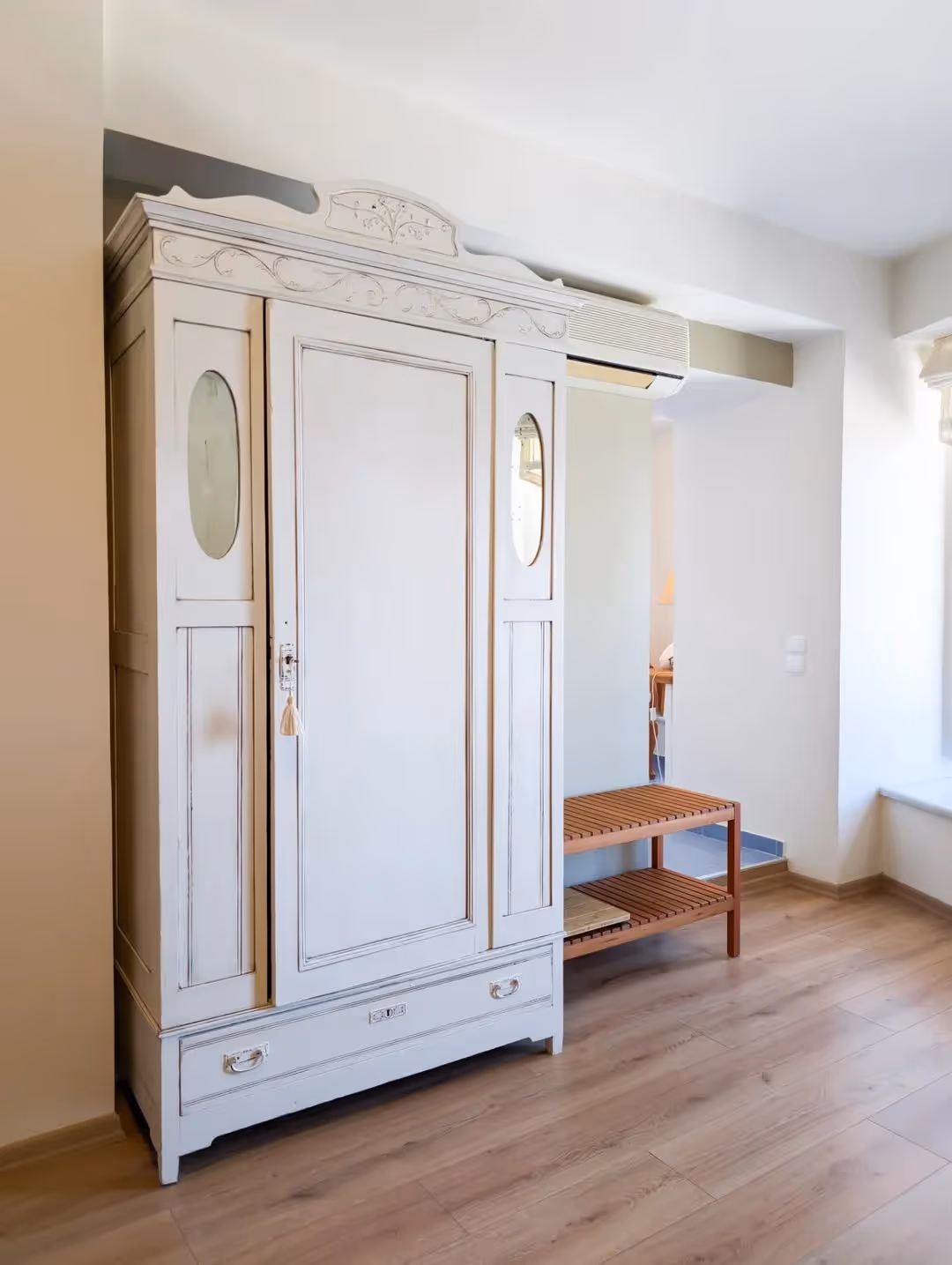 White wooden wardrobe with decorative carvings and two oval mirrors next to a wooden bench in a bright room.