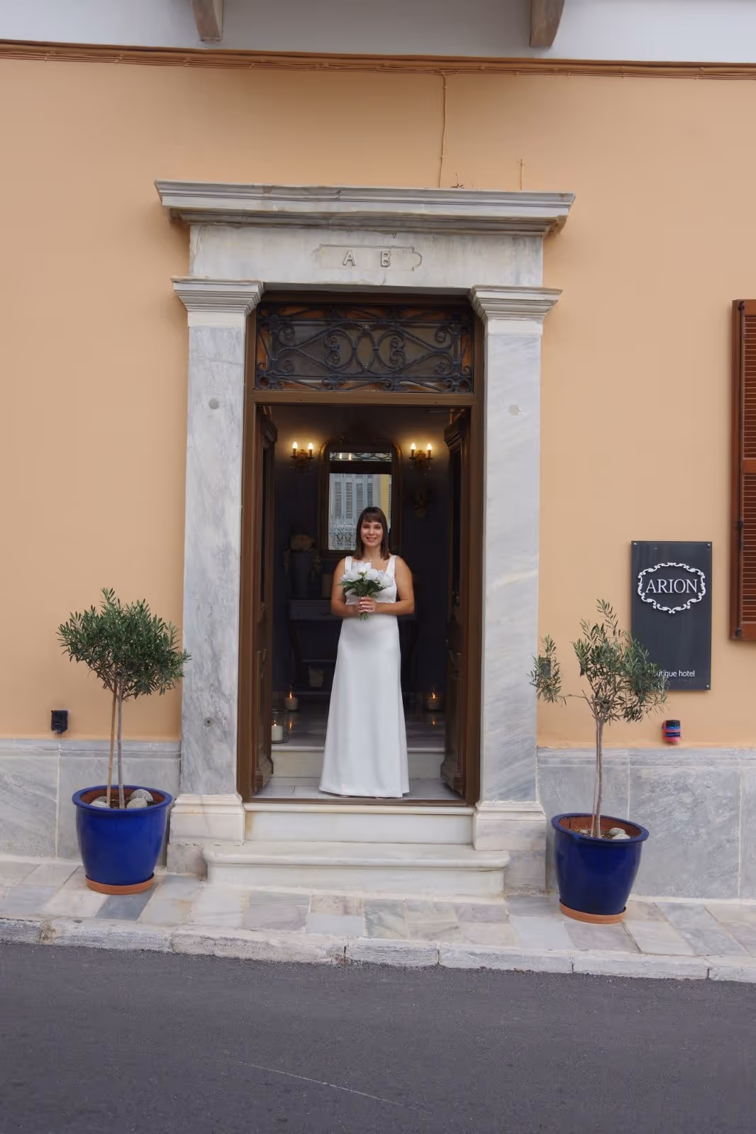 Woman in a white sleeveless dress holding a bouquet standing in a doorway of a building with potted plants on each side.