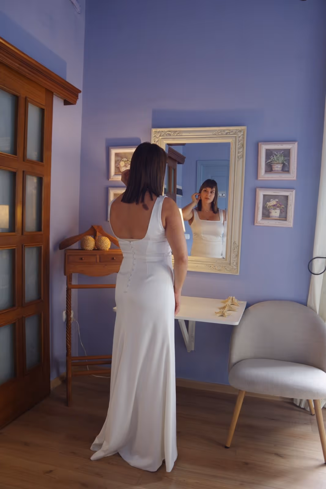 Woman in a white sleeveless dress standing in front of a wall mirror in a room with light purple walls.