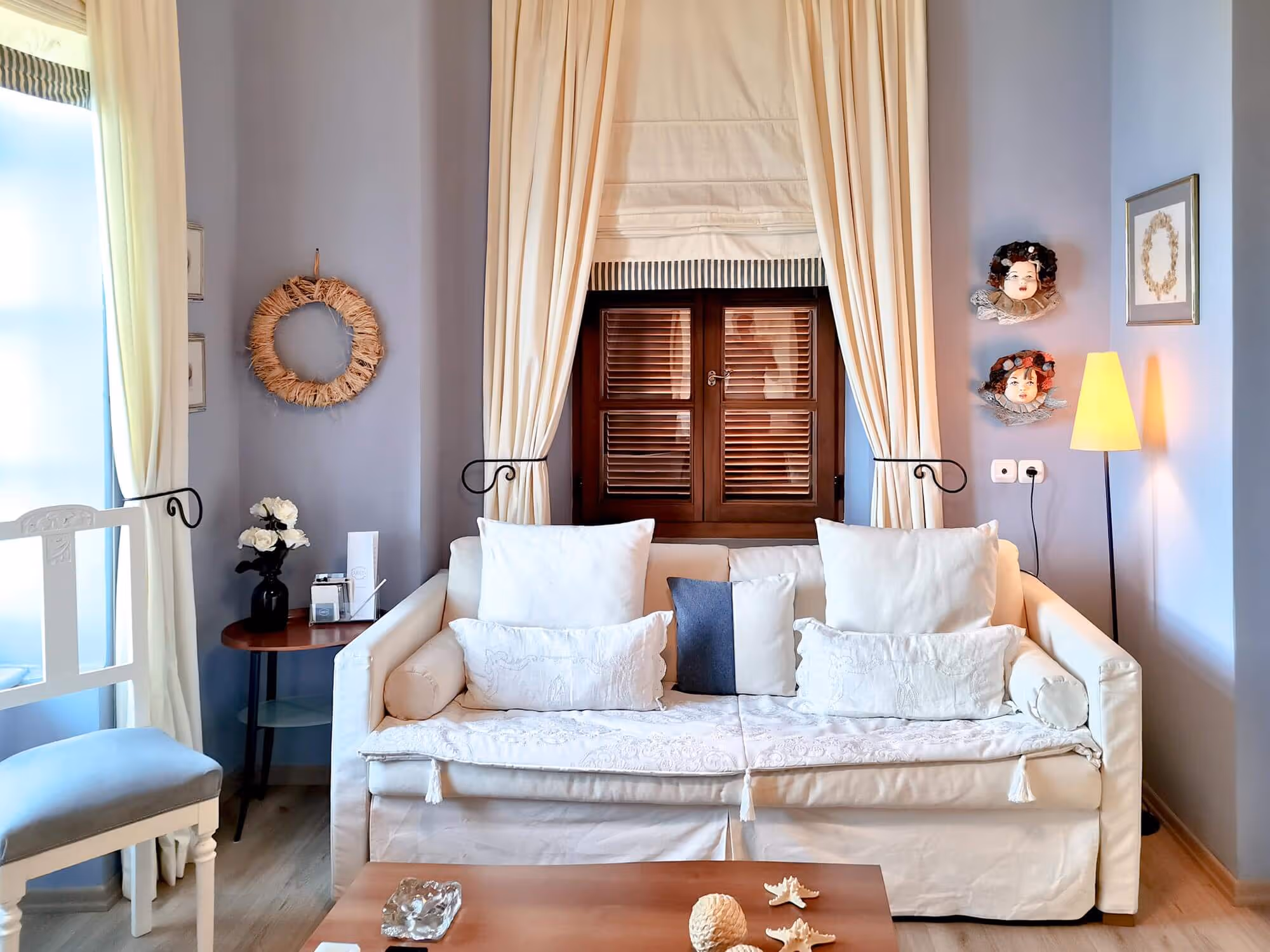 Cozy living room with white couch, decorative pillows, floral wreath, doll faces on wall, and floor lamp.