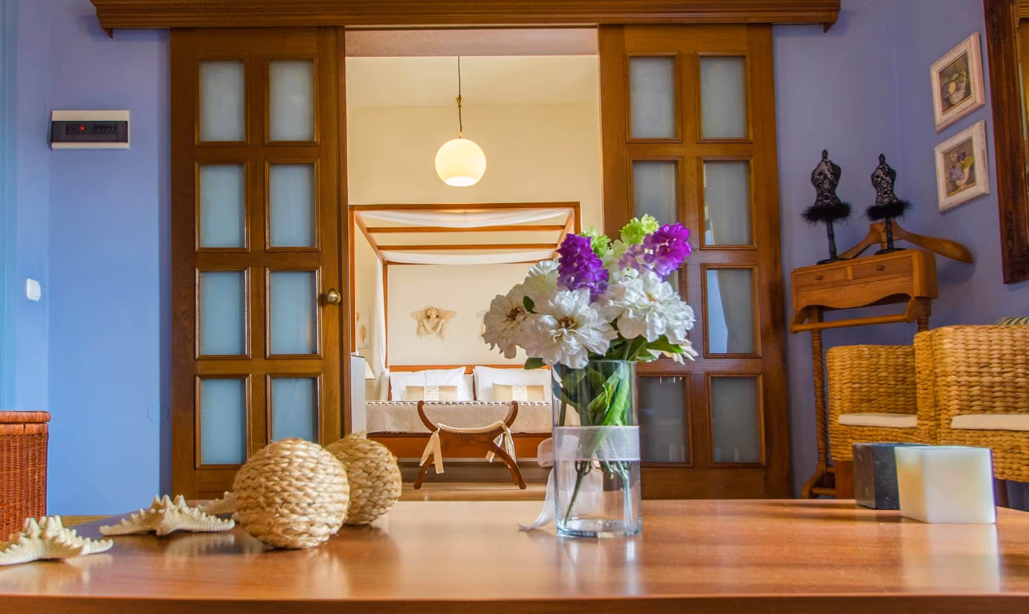 Cozy room with a wooden table foreground featuring a glass vase of white and purple flowers, wicker balls, and seashells, with a bed framed by wood sliding doors in the background.