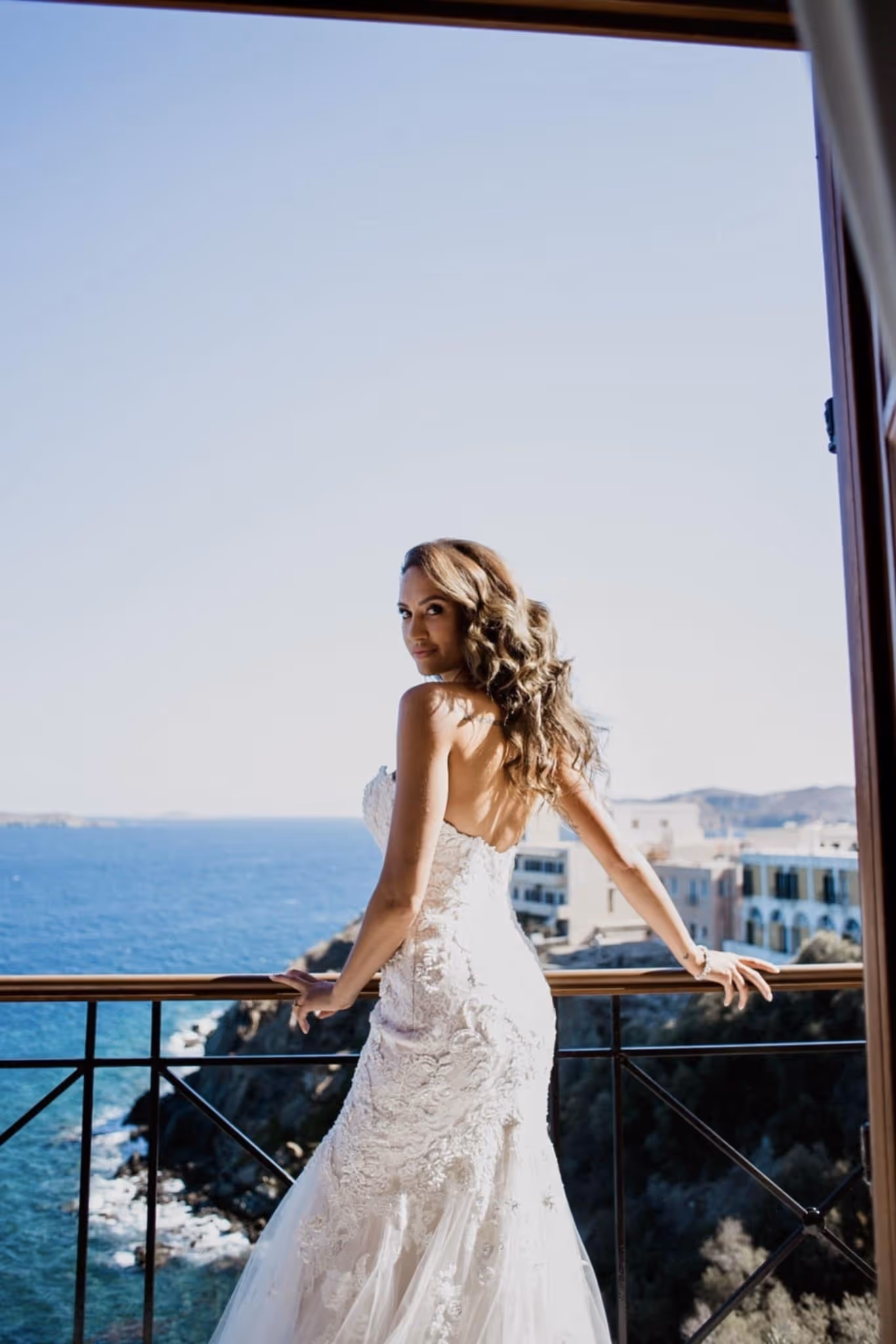 Bride in a strapless white lace wedding dress standing on a balcony overlooking the ocean and coastal buildings.