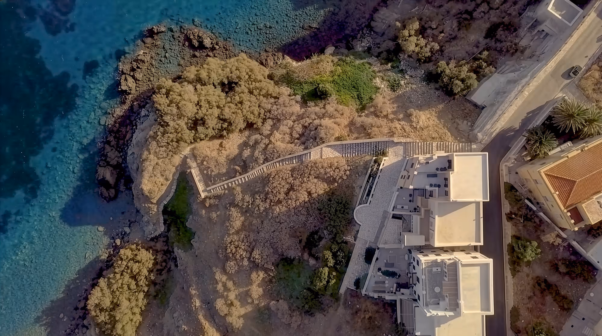 Aerial view of coastal buildings next to a rocky shoreline with clear blue water and a winding stairway path through shrubbery.