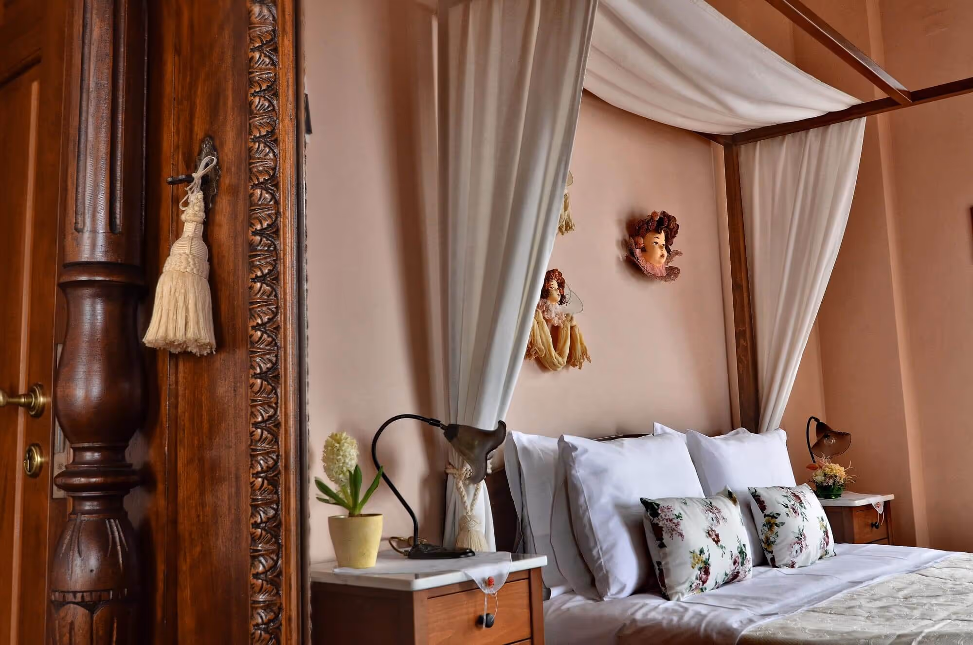 Cozy bedroom with a four-poster bed draped with white curtains, floral pillows, and wooden bedside tables with vintage lamps and decorative flowers.