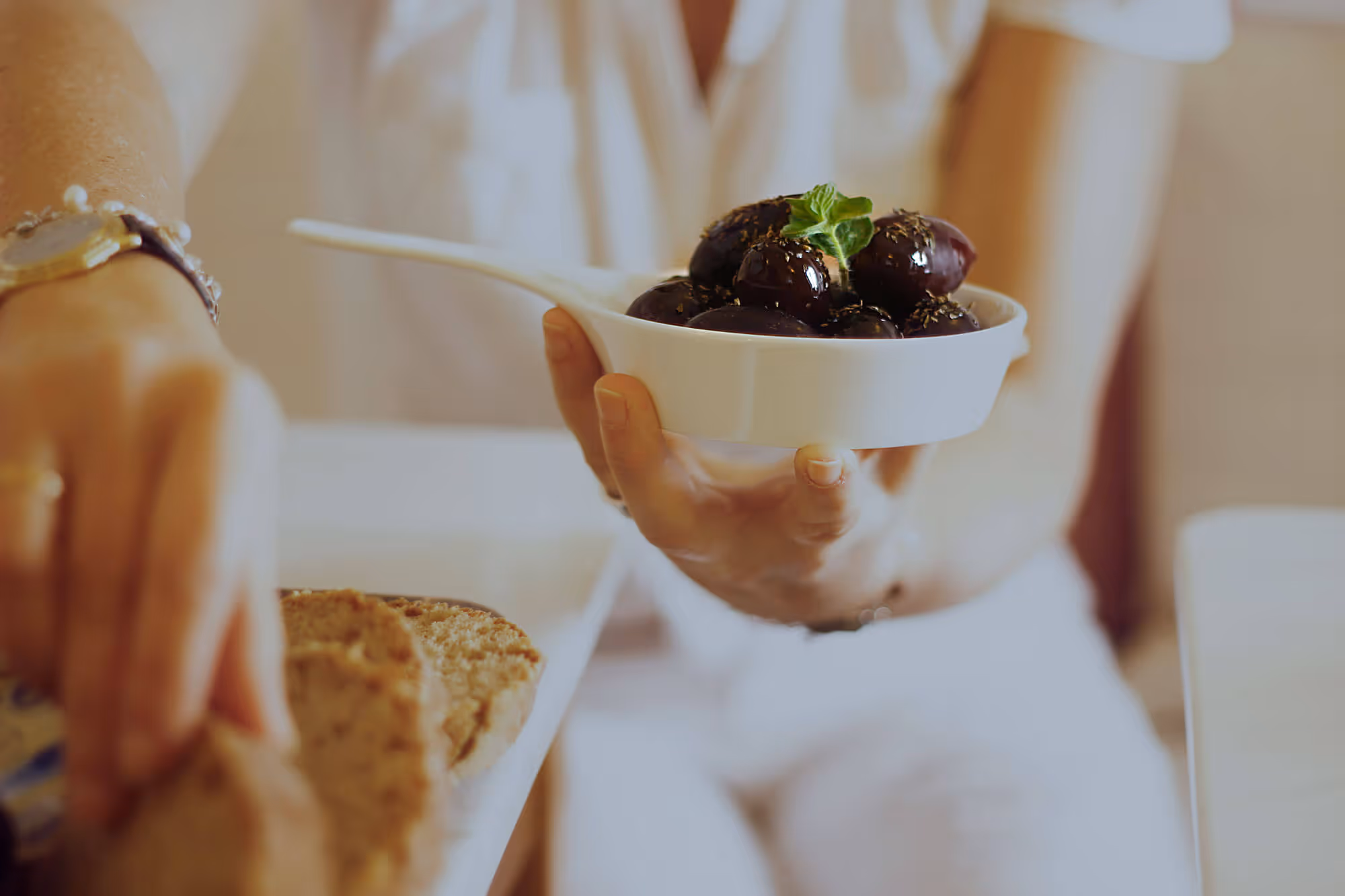 Hand holding a white dish filled with black olives garnished with herbs and a sprig of green leaf.