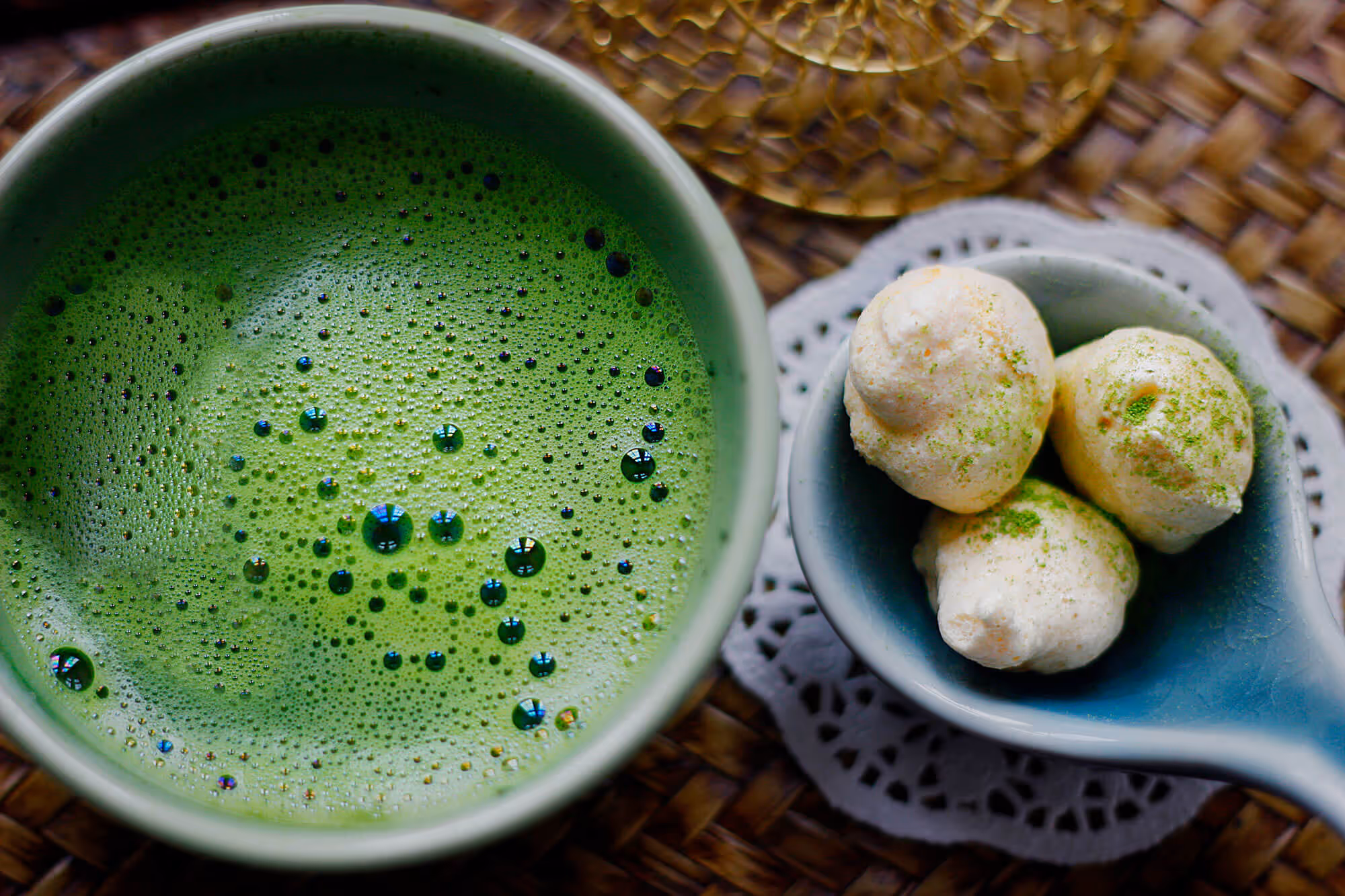 Close-up of a frothy green matcha tea in a cup beside a blue spoon holding three white meringue cookies dusted with green powder.