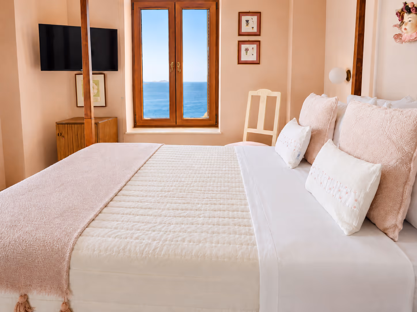 Bright bedroom with neatly made bed, pink pillows, a chair, TV, and ocean view through window.