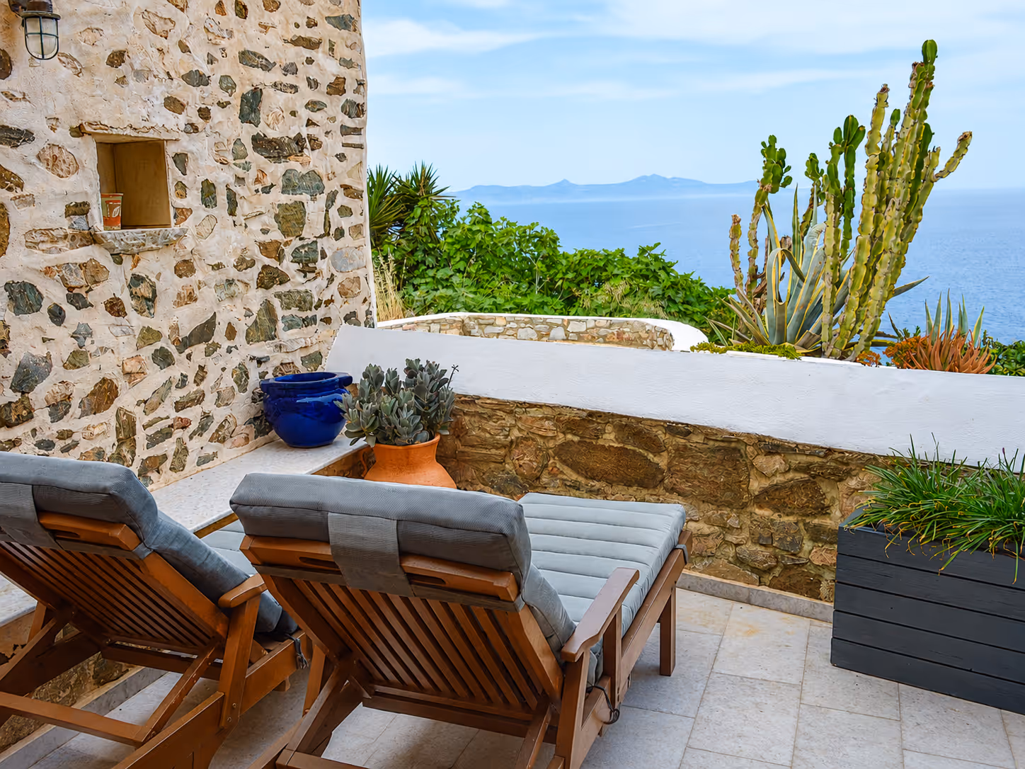Two cushioned wooden lounge chairs on a patio overlooking ocean and plants beside a stone wall.