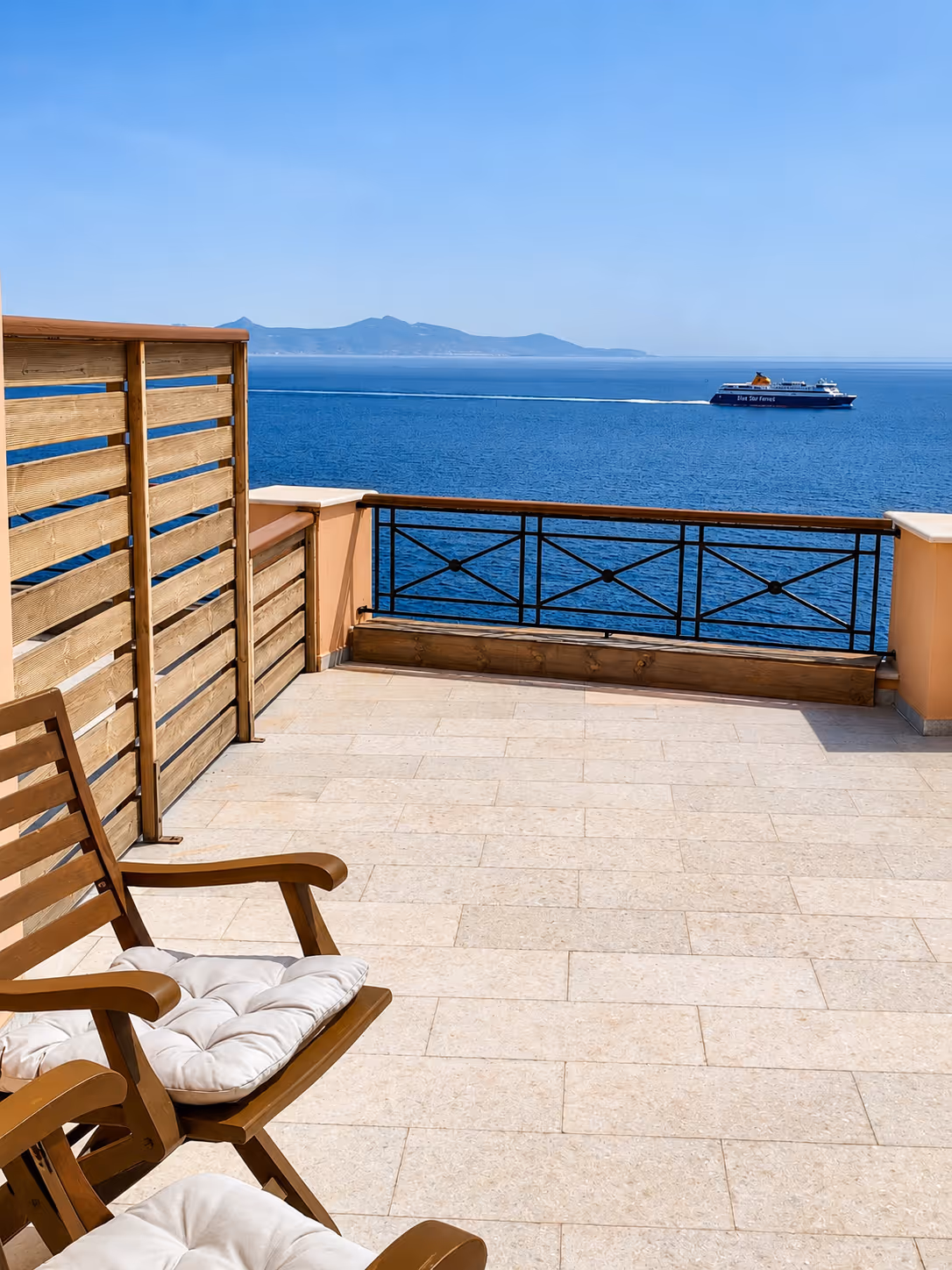 Balcony with cushioned wooden chairs overlooking blue sea and a ferry boat near distant mountains.