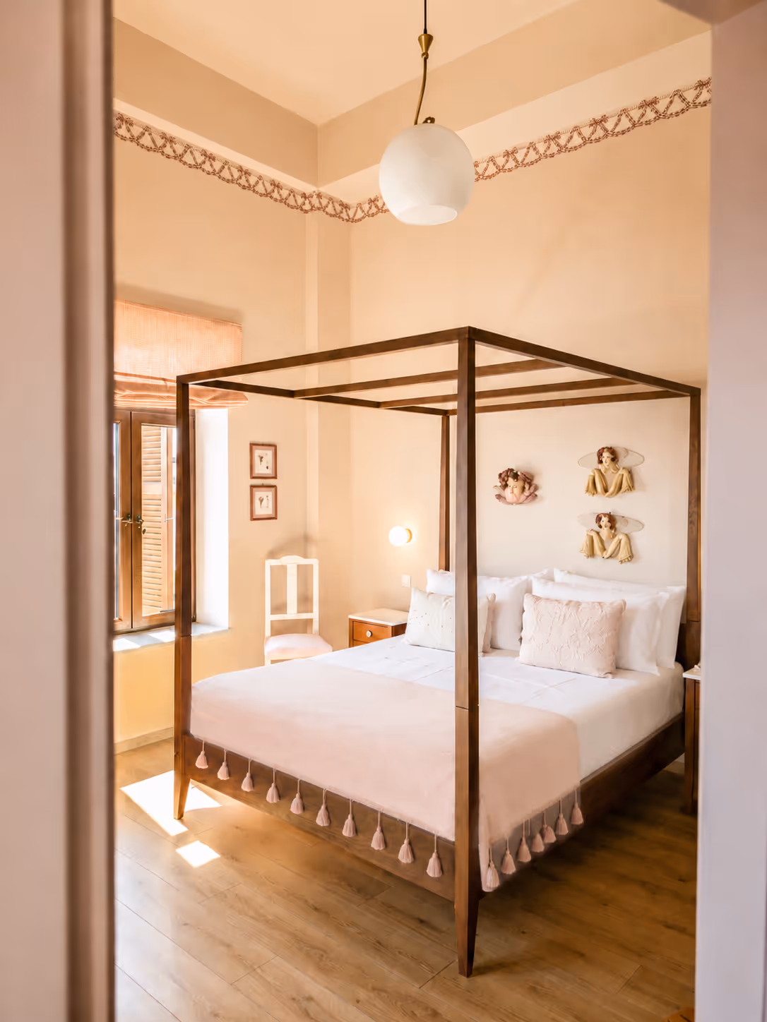 Sunlit bedroom with wooden four-poster bed, white linens, and decorative wall hangings of angels.