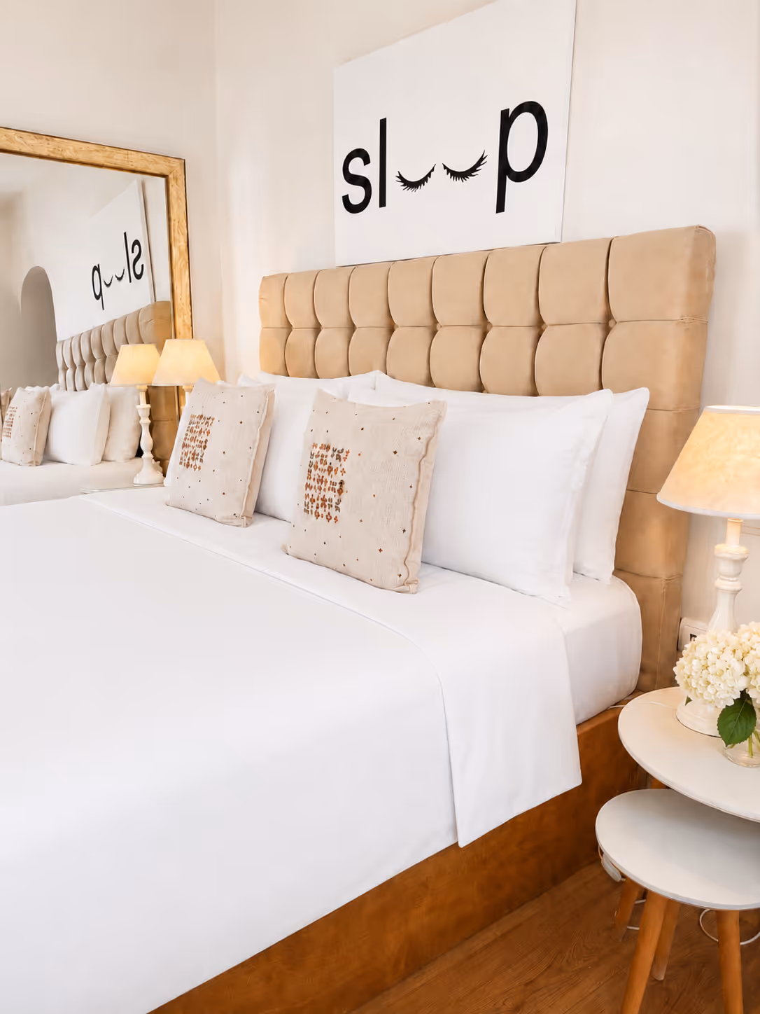 Neatly made bed with white linens, beige tufted headboard, decorative pillows, lamps, and a mirror.