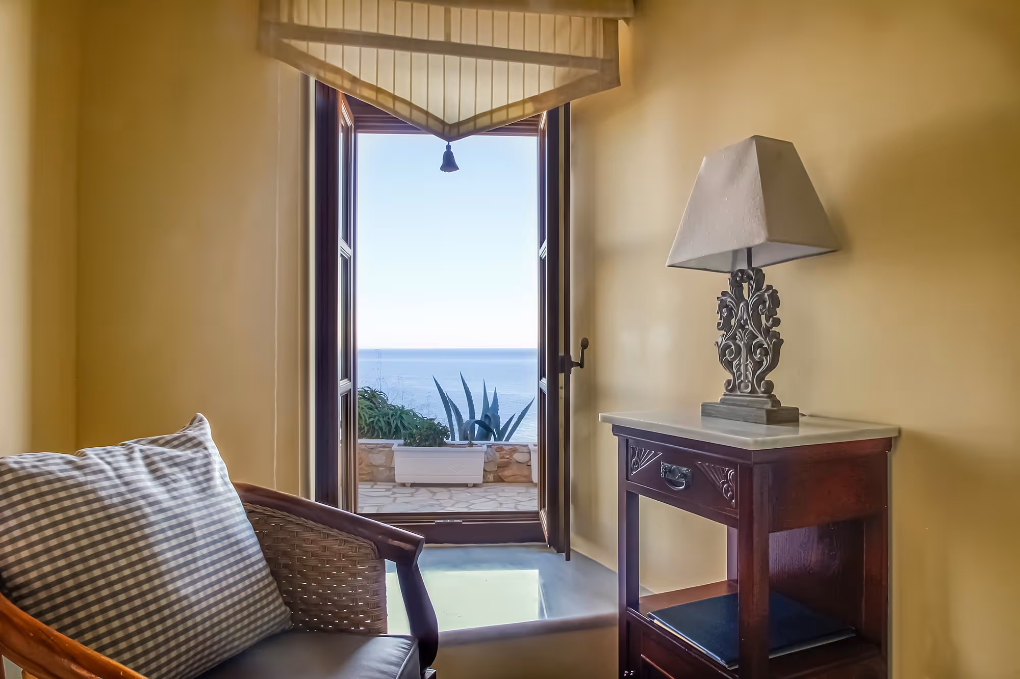 Cozy room with a wicker chair and pillow, an ornate dark wooden side table with a decorative lamp, and an open door revealing a sea view and potted plants outside.