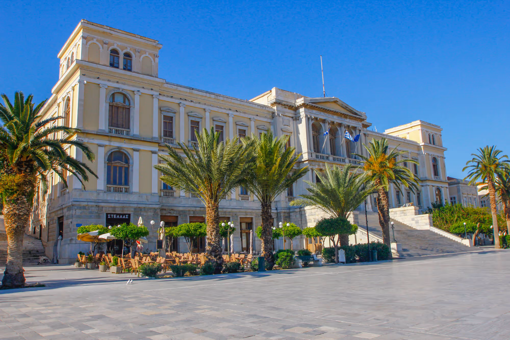 Large neoclassical building with Greek flags, palm trees, and outdoor seating under clear blue sky.