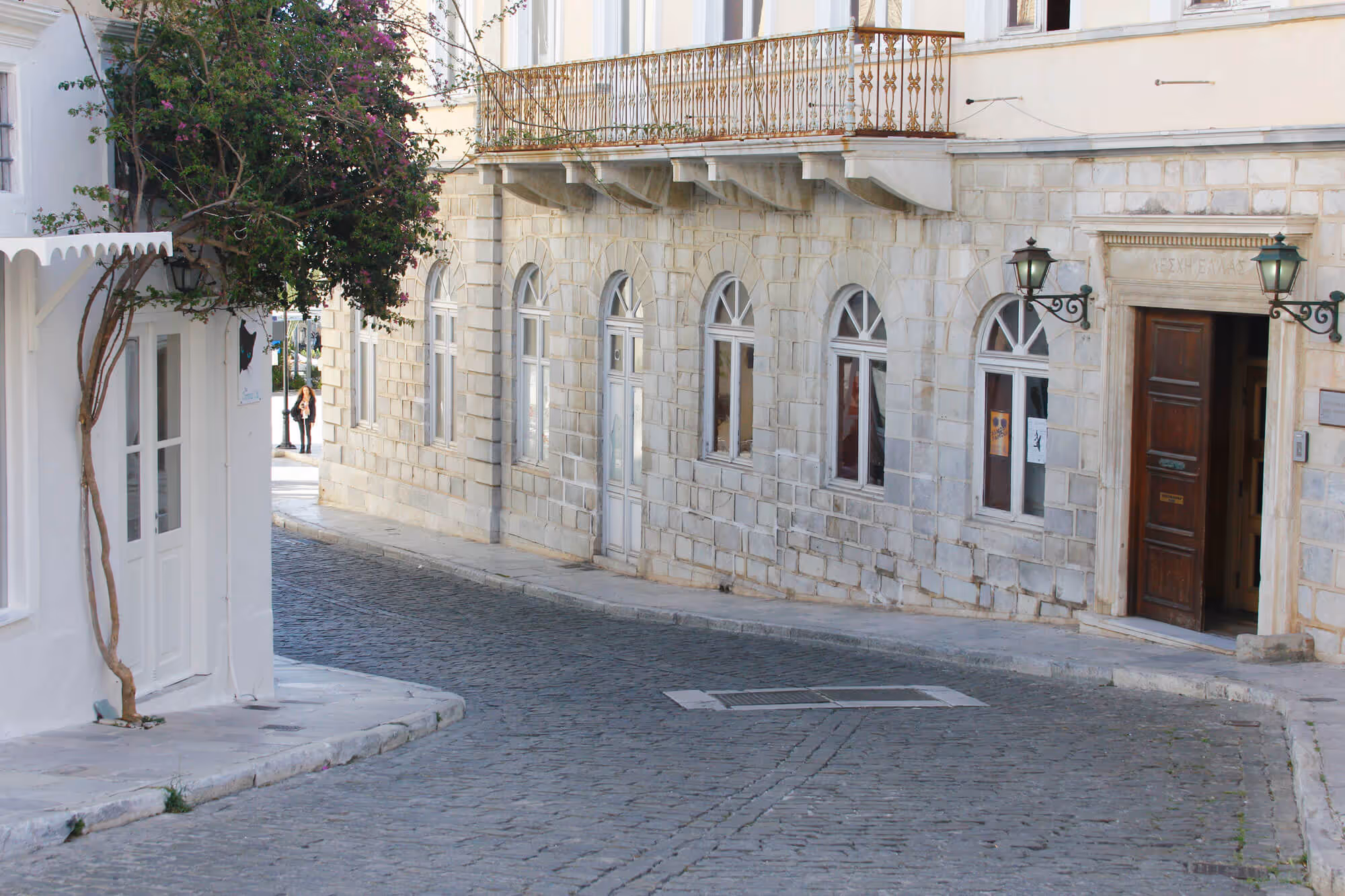 Curved cobblestone street beside white stone buildings with arched windows and a tree with purple flowers.