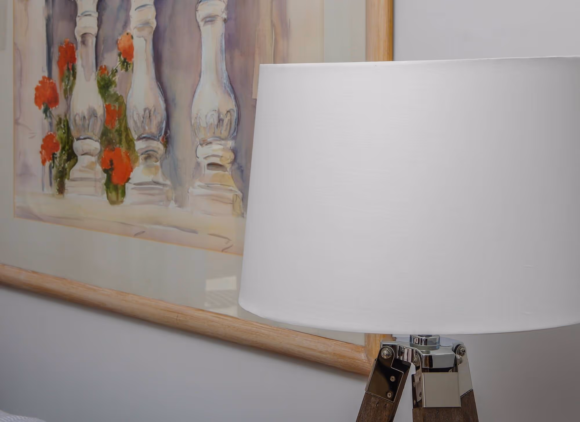 Modern tripod lamp with white shade beside a framed painting of stone balusters and red flowers.