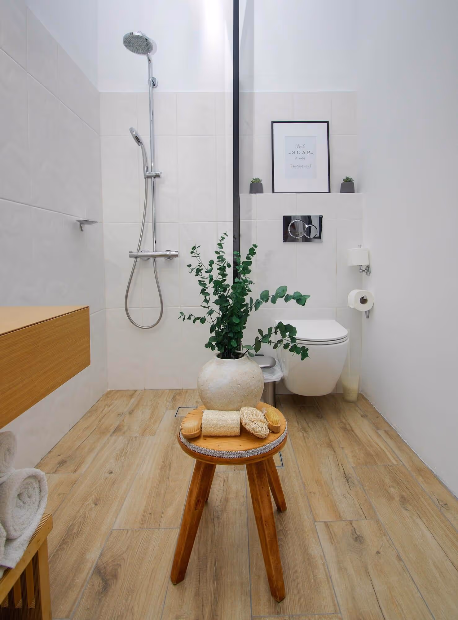 Modern minimalist bathroom with wooden floor, walk-in shower, wall-mounted toilet, and a small wooden stool holding a vase with green leaves and bath accessories.