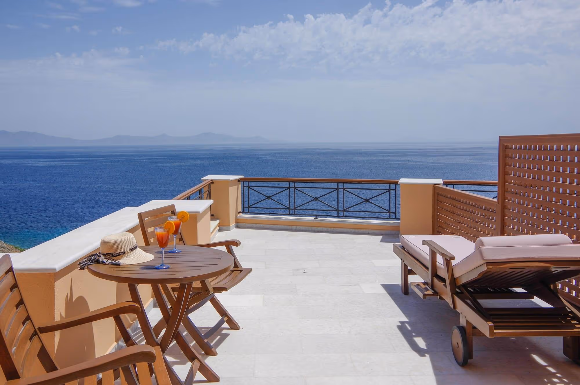 Terrace with wooden chairs, table with two cocktails and a sunhat, and a cushioned lounge chair overlooking a calm sea and distant mountains under a partly cloudy sky.