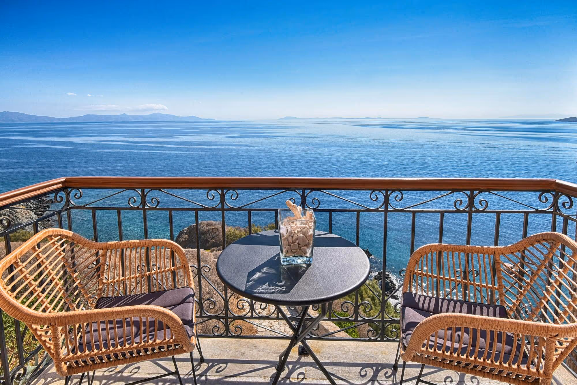 Balcony with two wicker chairs and a small round table overlooking a calm blue sea and distant mountains under a clear sky.