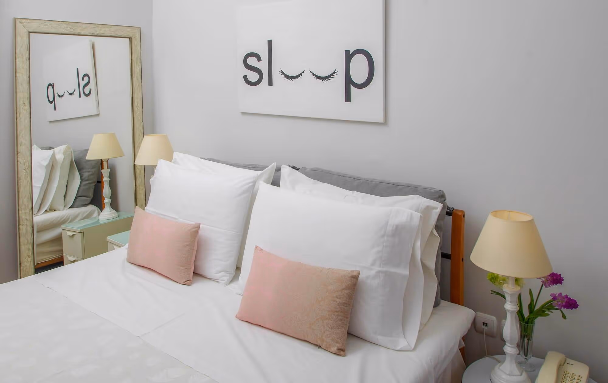 Bed with white and pink pillows, a bedside table with a lamp and flowers, and a wall art reading 'sleep' with eyelash graphics.