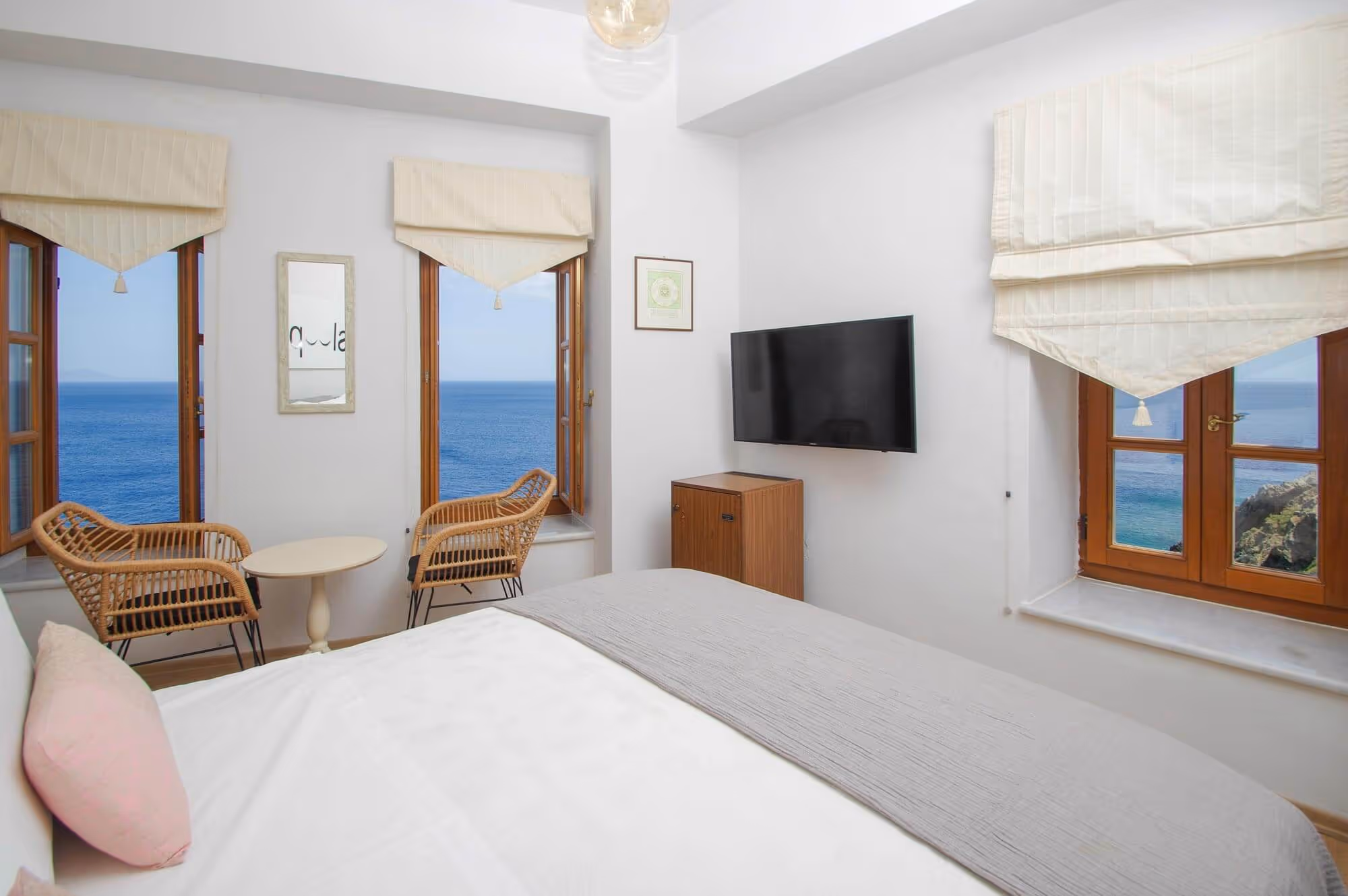 Bright bedroom with large bed, two wicker chairs, small table, wall-mounted TV, and windows overlooking a blue ocean.
